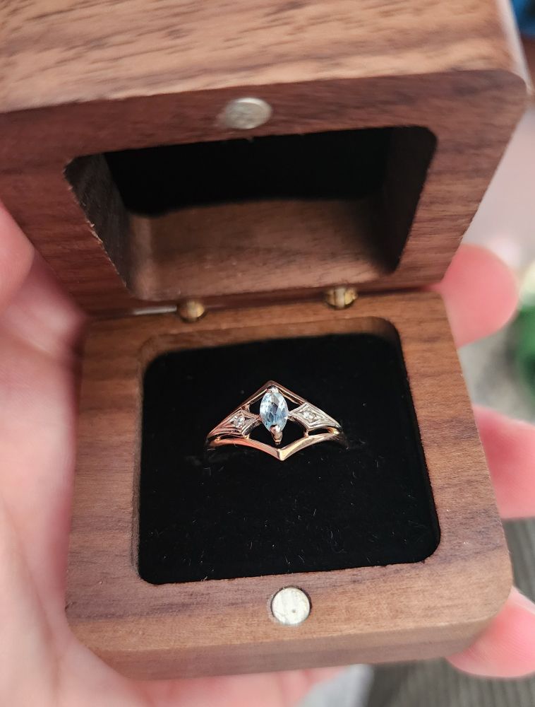 Art deco style ring with blue topaz for the gemstone, it sits in a black fabric insert in a dark brown, wooden box that closes with a magnet.