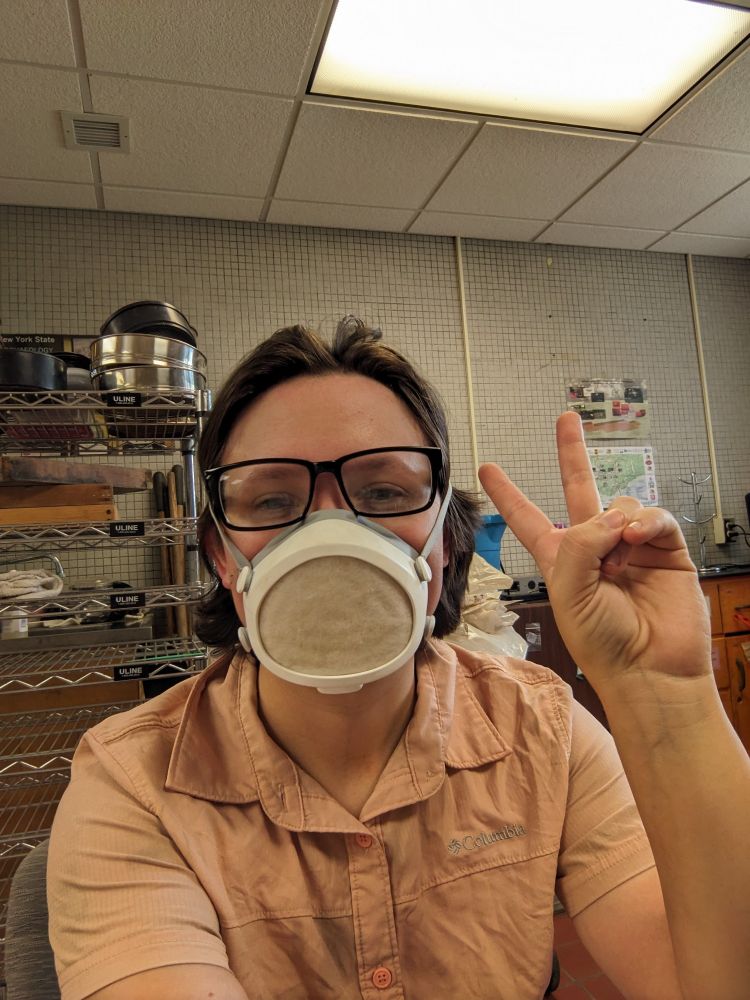 Kara wearing a pink button down field shirt and a flo mask, giving the peace sign. Signs of a smile crease their eyes.