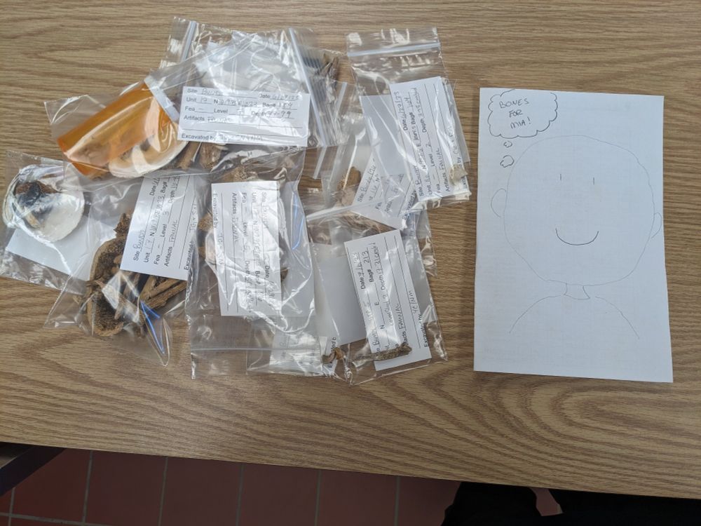On a table there is a pile of bags containing various faunal artifacts. To the right of the pile is a note which includes the sketch of a humans head and shoulders with a simple smiley face. The figure has a thought bubble that reads "BONES FOR MIA!"