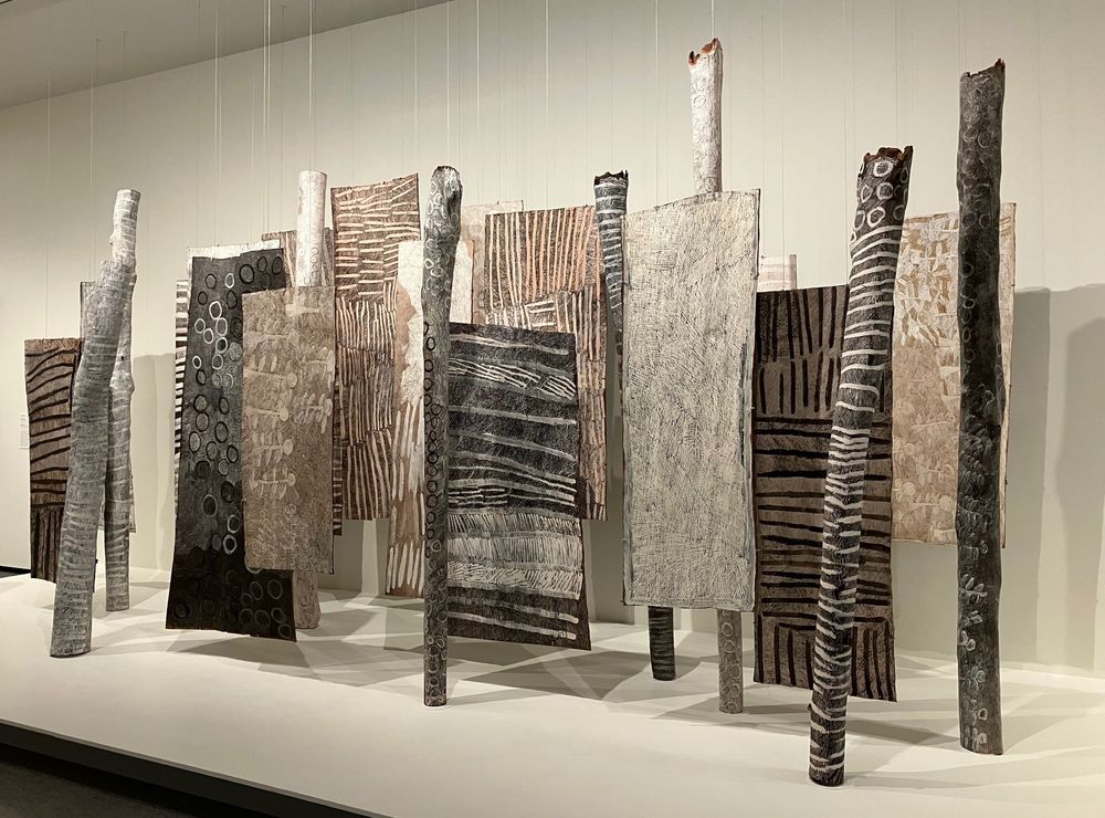 Gäna (Self) by Nyapanyapa Yunupinu

Nyapanyapa Yunupinu came from a line of important artists. As she recounted, "My father Munggurrawuy Yunupinu taught me how to paint. He said, When I am gone, you will follow behind me and paint too. Show the people — paint and work.'" After creating prints for over a decade, she began painting on bark in 2007, when she was in her 60s. This installation consists of 16 irregular bark paintings and nine larrakiti (memorial poles). Titled Gäna, meaning "self," this self-portrait reflects Yunupinu's intuitive style.