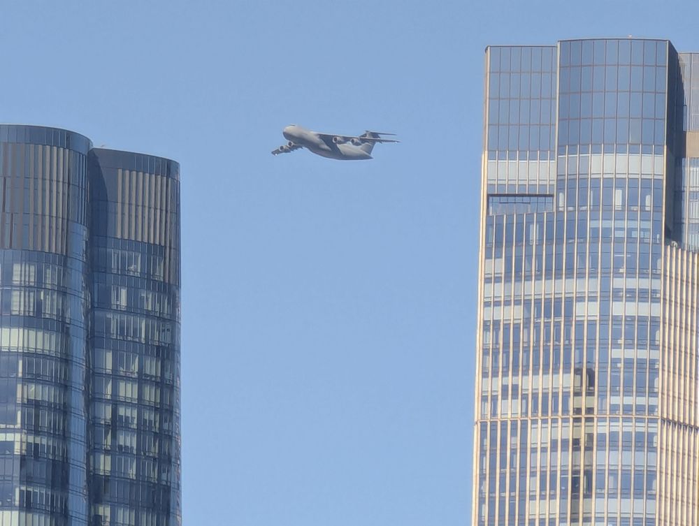 A large grey military cargo plane (C5 Galaxy, tail number 85-004) in a blue sky. In the foreground are two large buildings. 