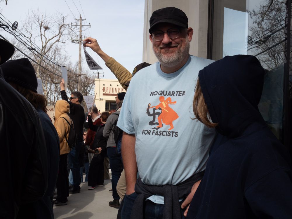 Photo from Tesla protest in Berkeley, CA on Saturday, Feb 15, 2025. Snapshot of a parent and child. The parent wears a shirt that reads "No quarter for fascists" and shows a person wrangling a snake in the shape of the fascist cross. Photo by Shana DeClercq