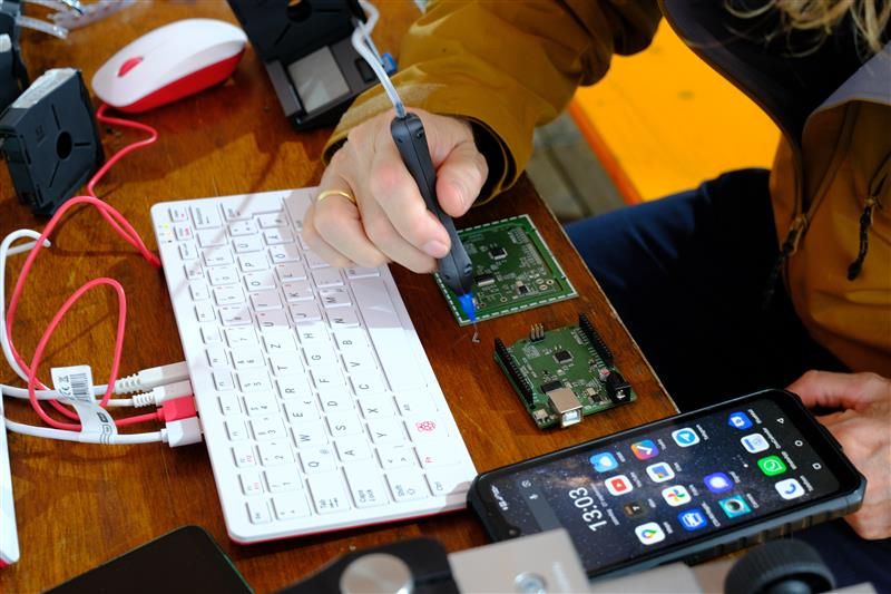 A close-up of a person using a handheld soldering tool on a green printed circuit board beside a Raspberry Pi keyboard and smartphone at the AISLER stand in Hall 2 at Maker Days 2025 in Eindhoven.