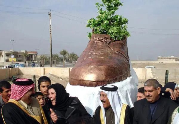 a bunch of iraqi people laughing and having a jovial time while standing around a large statue of the shoe that was thrown at george w bush. the shoe statue is bronze on a white pedestal and has a tree growing out of it, and is the size of a small car, rising up above the people in front of it.