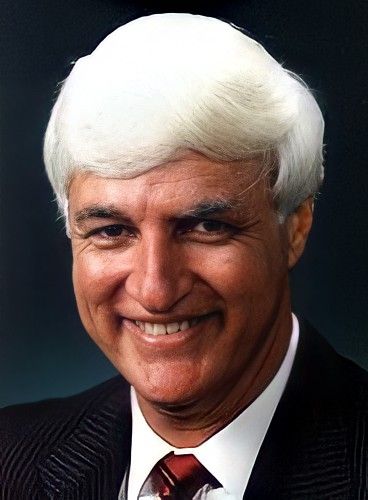 An image of Bob Katter, a man with what appears to be snow white ken doll sprayed on hair wearing a pin striped suit and smiling creepily, the archetype of "Queensland bloke politician"
