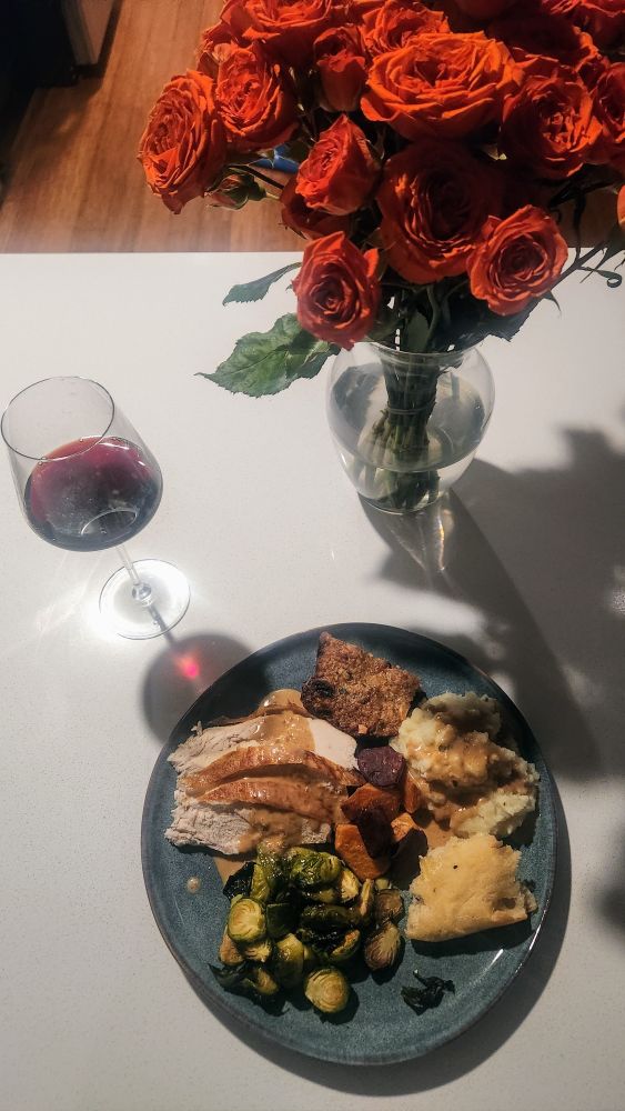 Bird's view of a blue dinner place with slices of breast covered with gravy, Brussels sprouts, bread, mash, a slice of dressing and a few purple and orange pieces of sweet potato in the middle. A stemmed glass of red wine stands on the top left, a vase with orange roses on the right. The light is hideous and rifle against the white countertop.