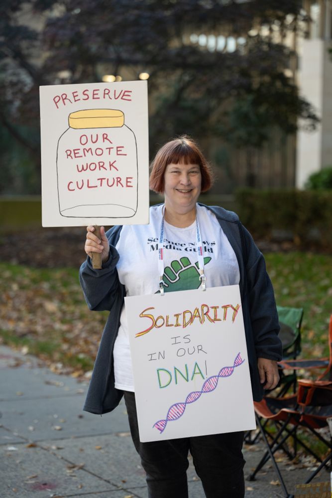 A Science News worker holds two signs, one in her right hand and another is on a lanyard around her neck. The one in her hand has a jar drawn on it and reads "preserve our remote work culture." The one around her neck reads "solidarity is in our DNA" and has a DNA helix drawn at the bottom.  