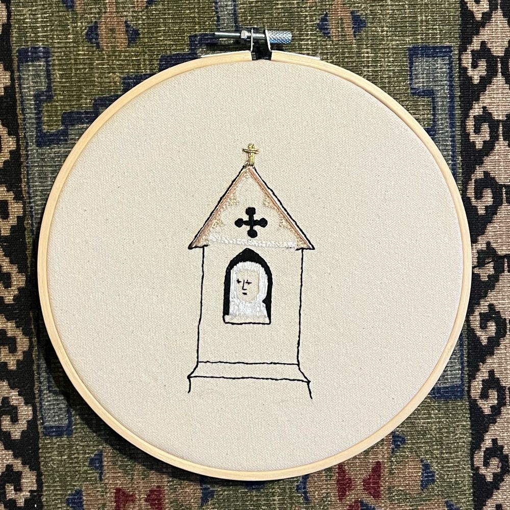 an embroidery hoop with an embroidered medieval picture of an anchoress looking out of a church window