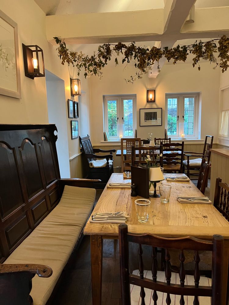 a pub with off-white walls and dark brown antique furniture laid out for sunday lunch, with dried hops hanging from the ceiling