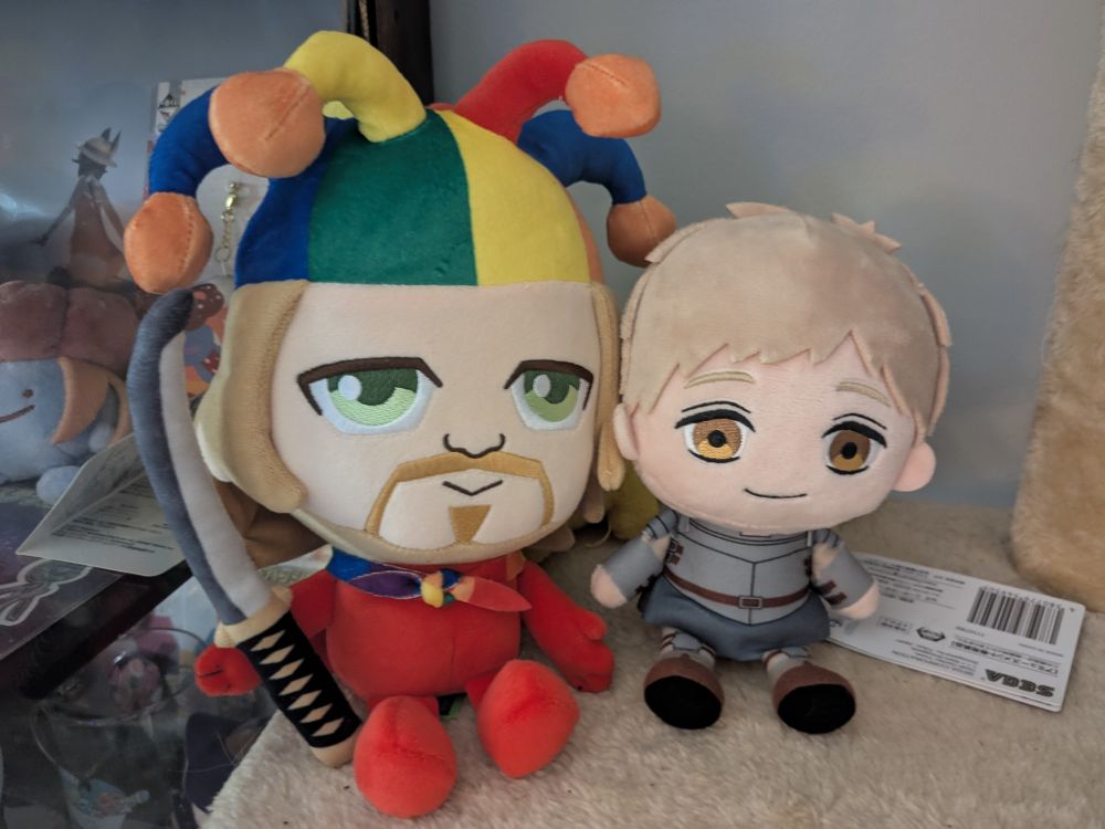 a plush of strawberry blond and eyebrowless YouTuber (jack! you have eyebrows!) in a colorful rainbow jester hat, wearing a rainbow flag cloak and a red suit, holding a katana, is sitting next to a plush of Laios Touden from Dungeon Meshi (a blond, golden-eyed guy wearing a suit of armor). Laios is smaller