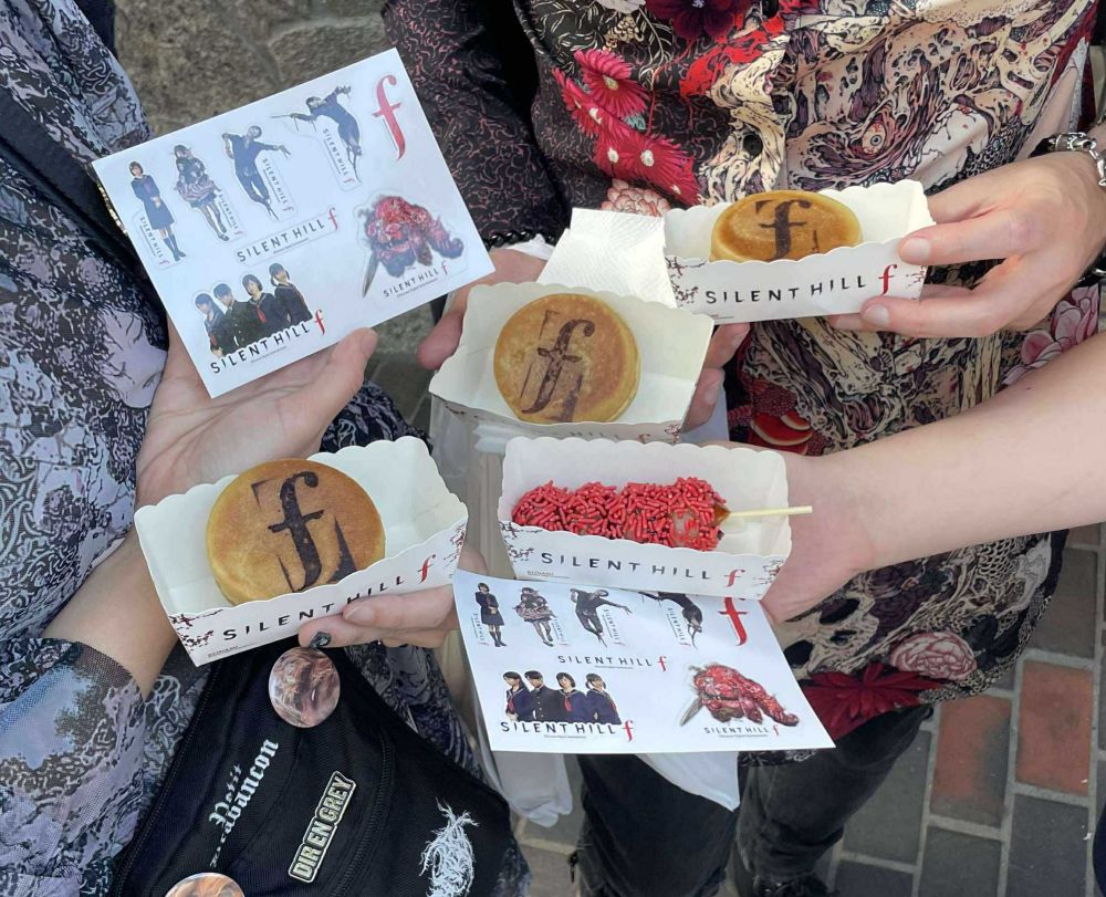 Snacks with the Silent Hill F logo and stickers of enemies and characters from the series held in people's hands. 