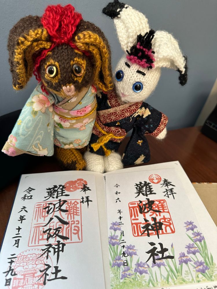 Fiddle & Jakebunny knit plushes posing over Fiddle’s Japan Temple & Shrine stamp book. The pages are open to the Goshuin stamps from Namba Yakasa Shrine & Namba Jinja Shrine. 