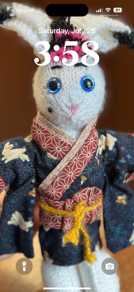 A locked phone screen, the time is in white font; the background image is a knitted white rabbit with blue eyes, it is wearing a blue kimono with red edges and a red obi belt. 