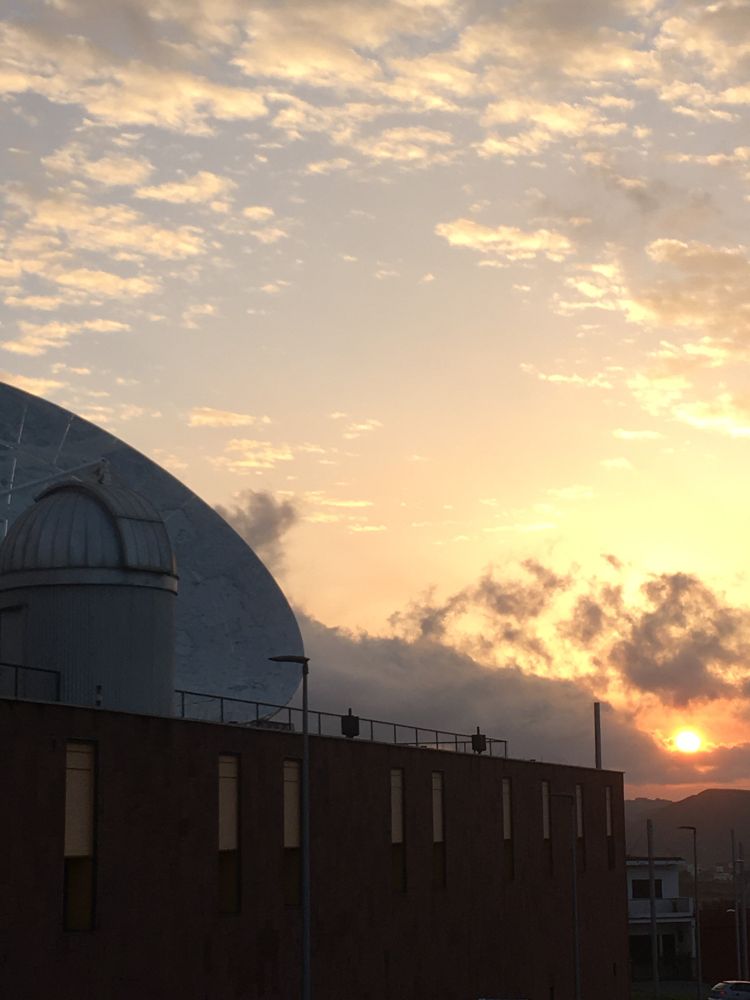 Fotografía de un amanecer. En primer plano la pared rojiza con ventanas del Museo de la Ciencia y el Cosmos, con su observatorio con cúpula y la antena del Radiotelescopio. Al fondo el sol anaranjado emergiendo de unas nubes sobre la cordillera de Anaga. Arriba el cielo, degradado de naranja a azul, con unas pocas nubes altas, como algodones.