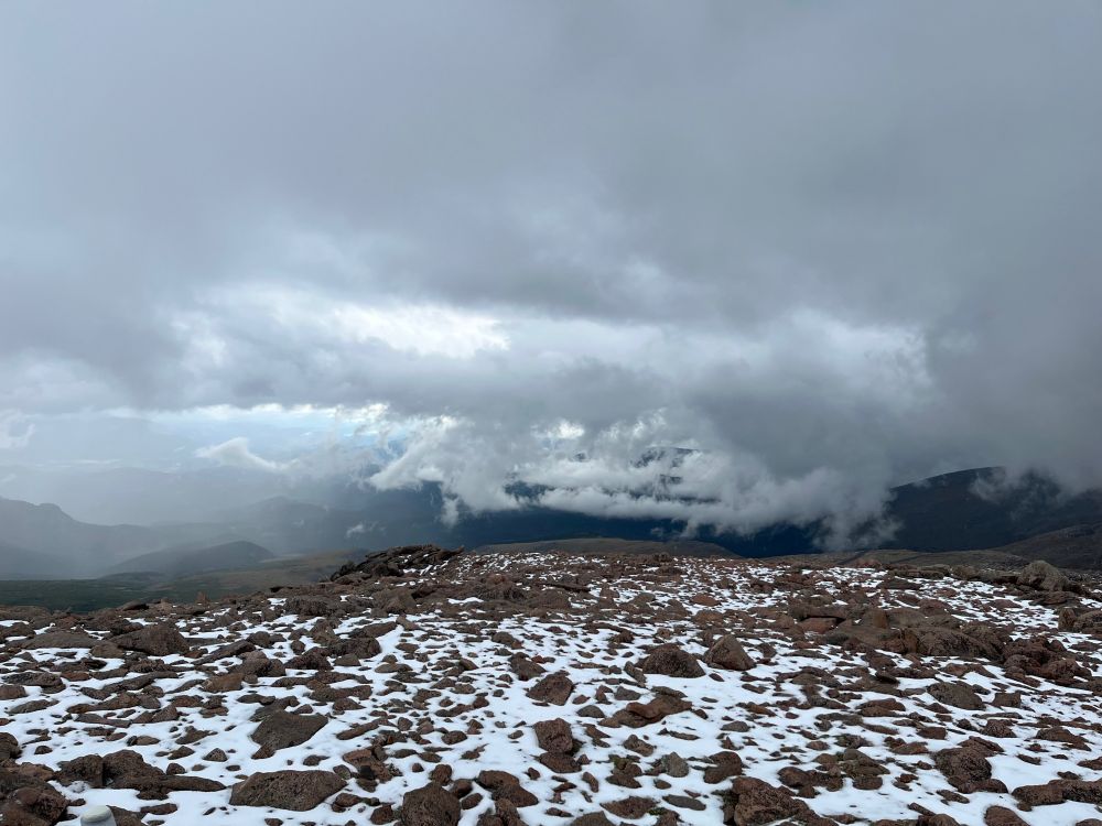 Mountain tundra scene with grey-brown rocks poking out of a thin layer of snow. Tattered dark grey clouds screen much of the distance, but at left a few faraway ridges are visible.