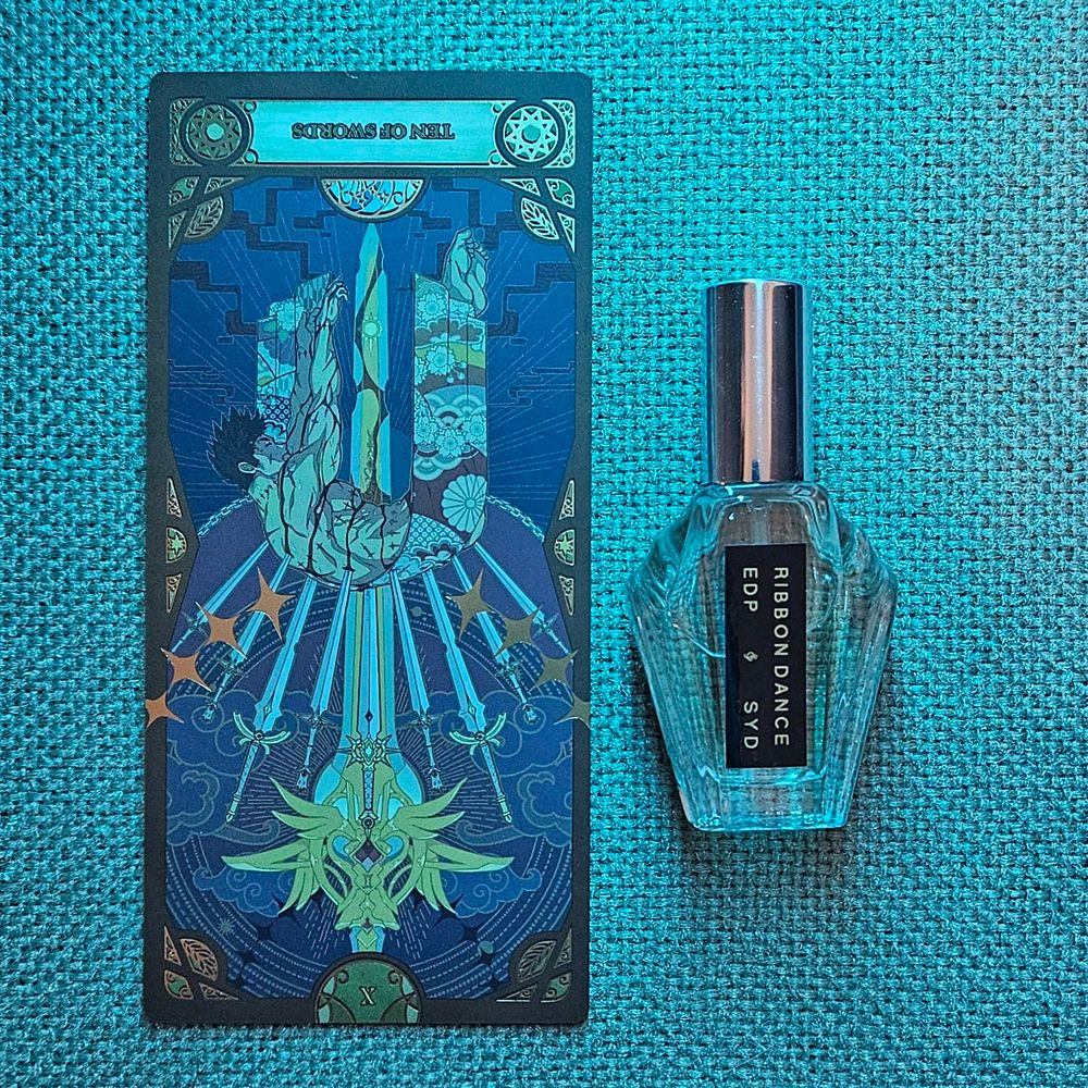 A reversed Ten of Swords tarot sits next to a bottle of Ribbon Dance by Syd Botanica