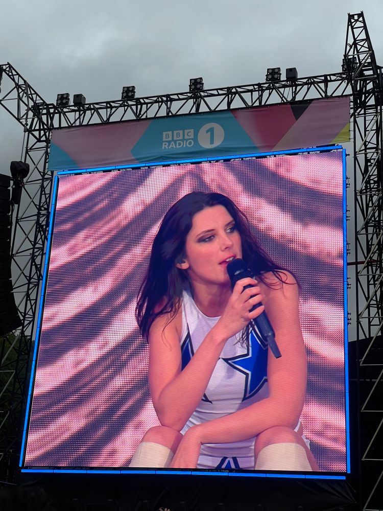 Ellie Roswell on stage at radio 1 big weekend photographed via the big screen to the side of stage where she is seen sitting down while she sings 