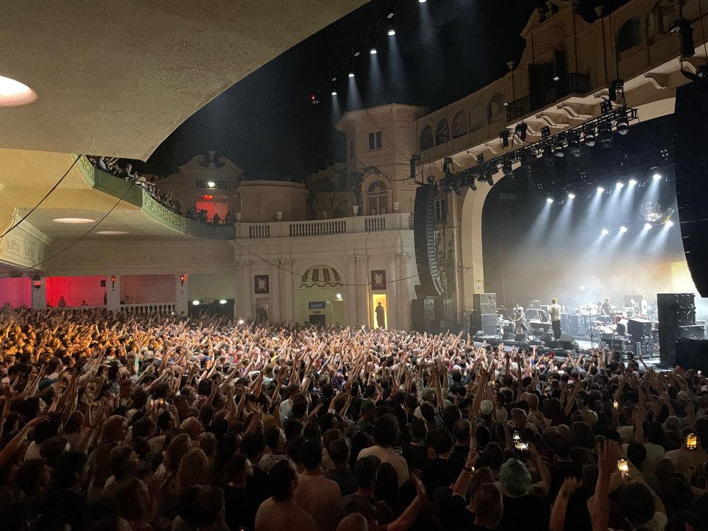 Big crowd at lcd soundsystem at Brixton academy everyone has hands in their and is lit by lights coming down from the stage 