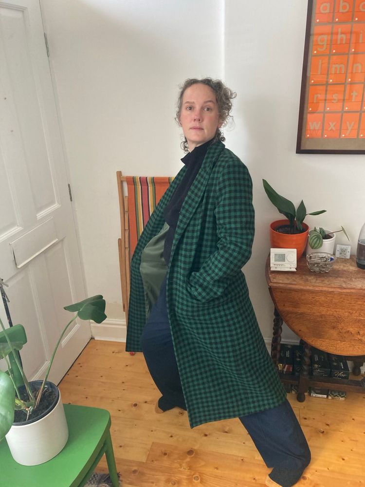 Middle aged autistic chaos in human form wearing wide legged trousers and a bottle green and black checked overcoat,. doing what looks like a side lunge, for reasons I cannot fathom. There are definitely not several boxes of wine under the table behind her.