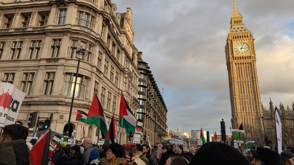 Crowds of people in London for a rally for Palestine. Many are carrying Palestinian flags. In the background, the tower of Big Ben stands golden in the evening light. 