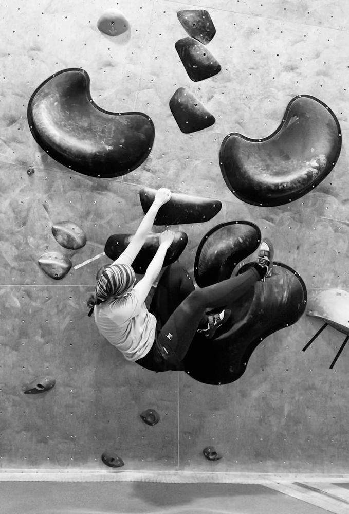 Ich beim Bouldern (monochromes Foto):
Ich hänge mit ausgestreckten Armen seitlich an zwei größeren Griffen, die dicht übereinander an die Wand geschraubt sind. Der linke Fuß steht auf einem riesigen Tritt deutlich oberhalb vom Po, das Knie ist maximal gebeugt. Das rechte Bein strecke ich mit leicht gebeugtem Knie schräg nach oben in die Luft. Der Po ist der tiefste Punkt und ich schaue in Richtung meiner Knie. Ich trage eine geringelte Mütze, ein helles T-Shirt und eine dunkle Hose.