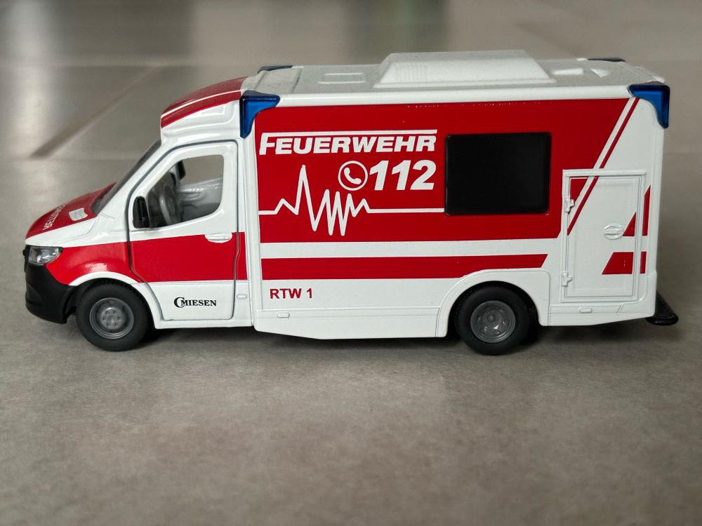 Ein weiterer Spielzeugrettungswagen mit rot und weißer Lackierung. Auf dessen Seite ist ein Quatsch-EKG zu sehen, es besteht einfach aus ein paar zufälligen Zacken.