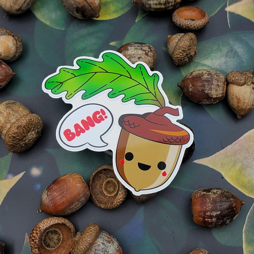 A die-cut sticker featuring a kawaii-style acorn character with a smiling face, rosy cheeks, and a brown cap, topped with a green oak leaf and saying 'BANG!' in a speech bubble. The sticker is surrounded by real acorns on a surface with a dark leaf print.