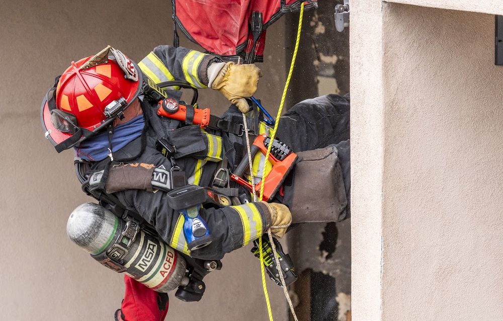 Firefighter in red helmet and full turnout gear with SCBA tank rappels down the exterior wall of a building using rope rescue techniques, practicing emergency evacuation.