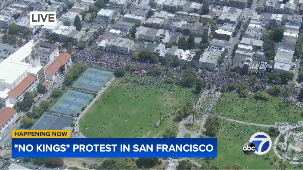 Aerial helicopter view from ABC 7 news broadcast showing a large crowd gathered in a San Francisco park and surrounding streets for a 'No Kings' protest. The image shows thousands of people filling a green park area with tennis courts visible, as well as spilling onto adjacent residential streets lined with multi-story buildings. The scene captures the scale of the demonstration from above. A news broadcast overlay indicates this is live coverage, with 'HAPPENING NOW' and '"NO KINGS" PROTEST IN SAN FRANCISCO' text, along with the ABC 7 logo.