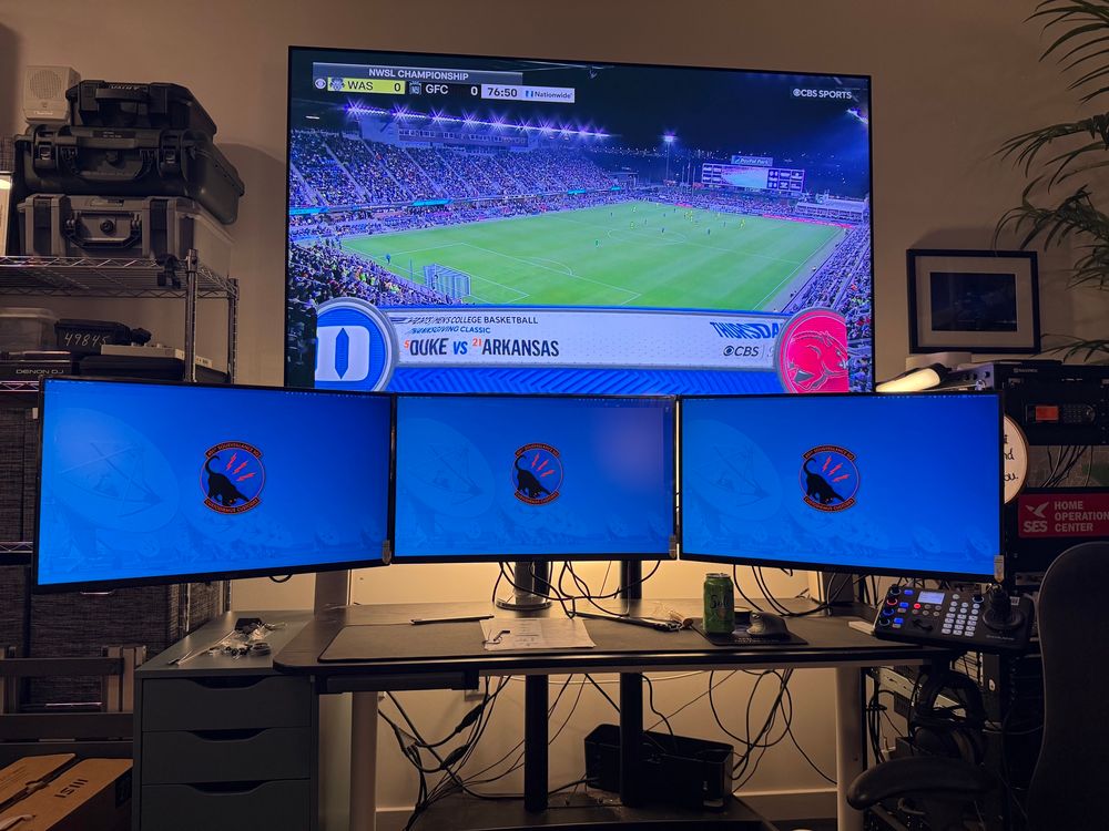 photo of a messy triple monitor home office setup with the monitors on a mount that places them too far over the front of the desk

a large TV over the desk setup has NWSL soccer on