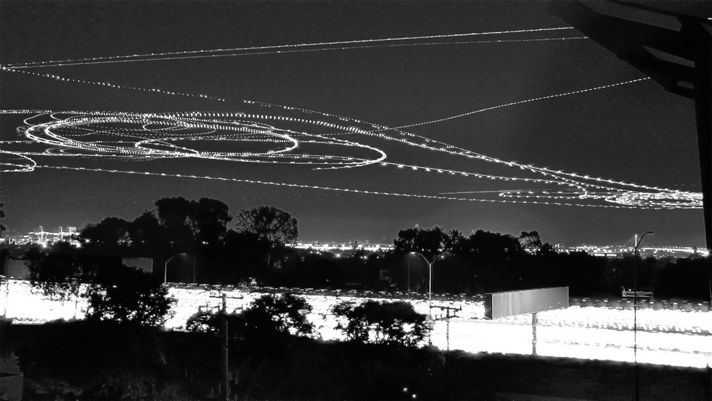 A black and white long-exposure photograph showing aircraft light trails creating curved streaks and spirals across the night sky. The trails appear as continuous white lines of varying intensity, capturing the flight paths of multiple planes over time. Below, silhouettes of trees, buildings, and street lights are visible along the urban horizon.