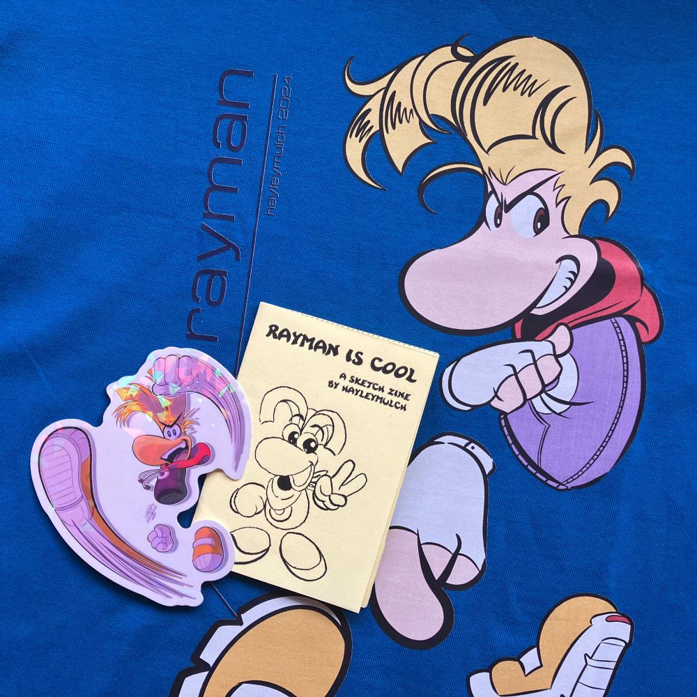Photo of some Rayman fan merch by Hayley Mulch. It features a close-up of a blue Rayman t-shirt along with a holographic Rayman sticker and a small, yellow zine titled "Rayman Is Cool" placed on top of the shirt, partially covering the graphic.