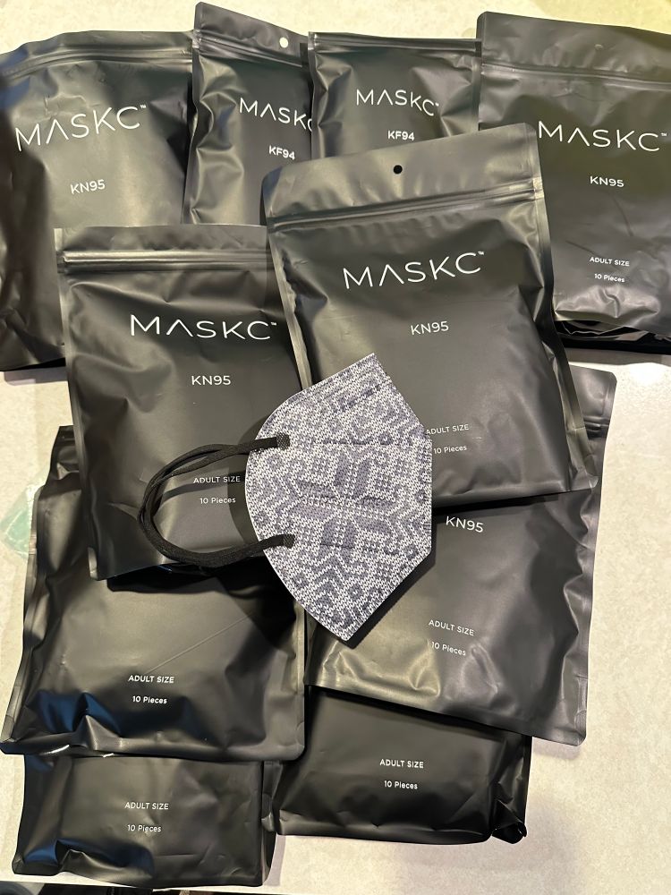 An assortment of black resealable packages labeled ‘MASKC’ containing KN95 and KF94 adult-size masks, with a gray face mask featuring a classic winter knit pattern printed on its surface displayed in the foreground. Each package indicates a count of 10 masks.