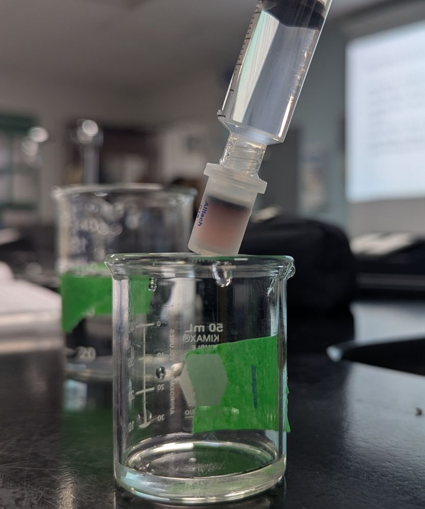A beaker with some clear fluid inside sits underneath a syringe attached to a chromatography separation column. The inside of the column is red/pink with a dark blue line at the top. 