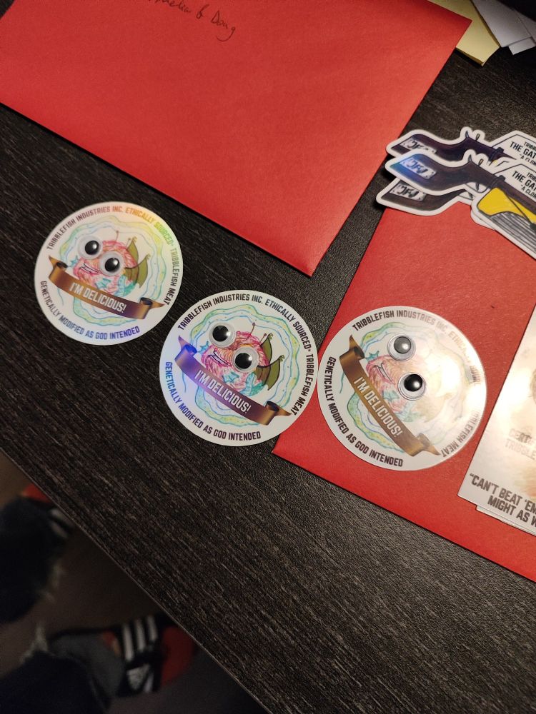 3 identical circular stickers showing a fuzzy flying fish with a long tentacle that goes around the circle of the sticker.

There are googly eyes on the fish, a banner that reads "I'm delicious!"
Around the edge of the sticker there is text that reads "tribblefish industries Inc  ethically sourced* tribblefish meat"
And "genetically modified as god intended"