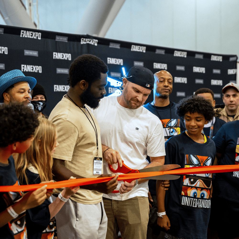 Image of a rope cut on the red carpet at FAN EXPO Boston with Josh Olumide and Liam Hendricks with a group of kids surrounding them watching and smiling.
