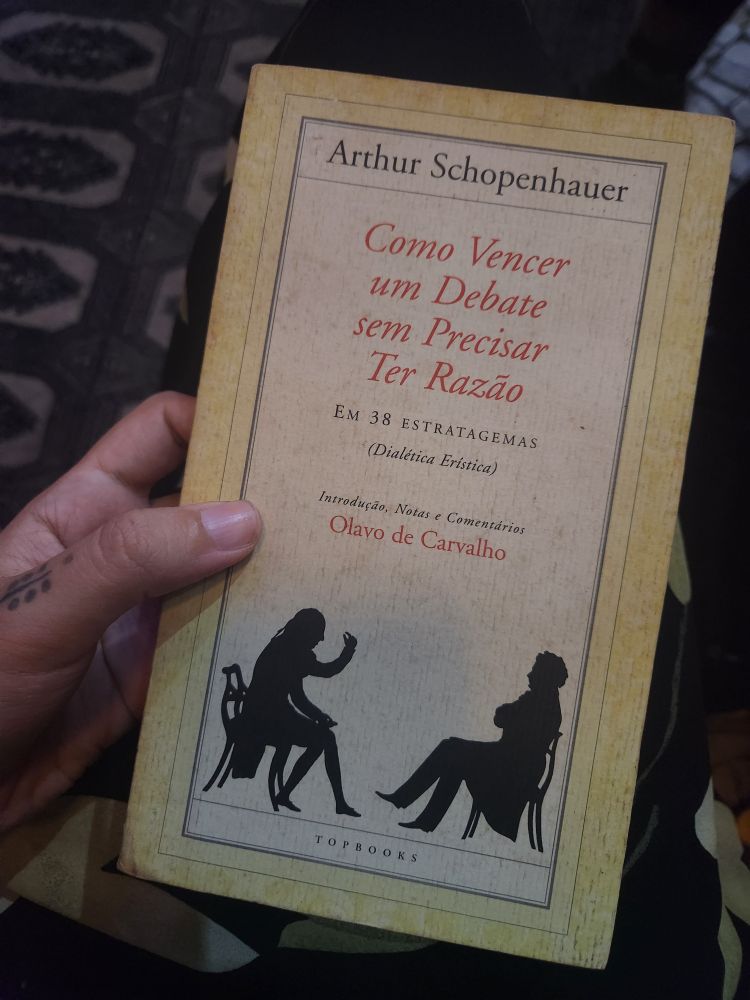 minha mão segurando o livro "como vencer um debate sem eoeciaae ter razão" de arthur schopenhauer com introdução, notas e comentários do defunto olavo de carvalho