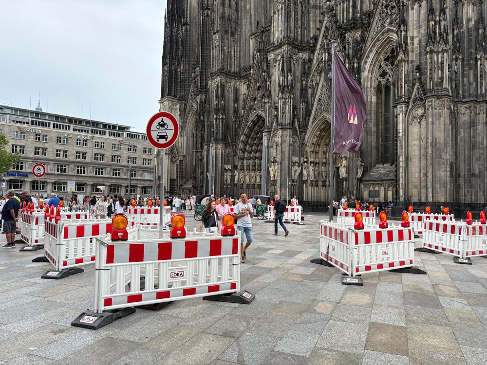 Westfassade des Kölner Doms mit der Domplatte davor, auf der eine Menge rot-weißer Kunststoffabsperrungen in Dreiecken aufgebaut sind sowie ein Schild, dass die Fläche nicht von Motorrädern und PKW befahren werden darf. Hintergrund ist eine schadhafte Säule im Parkhaus darunter. 