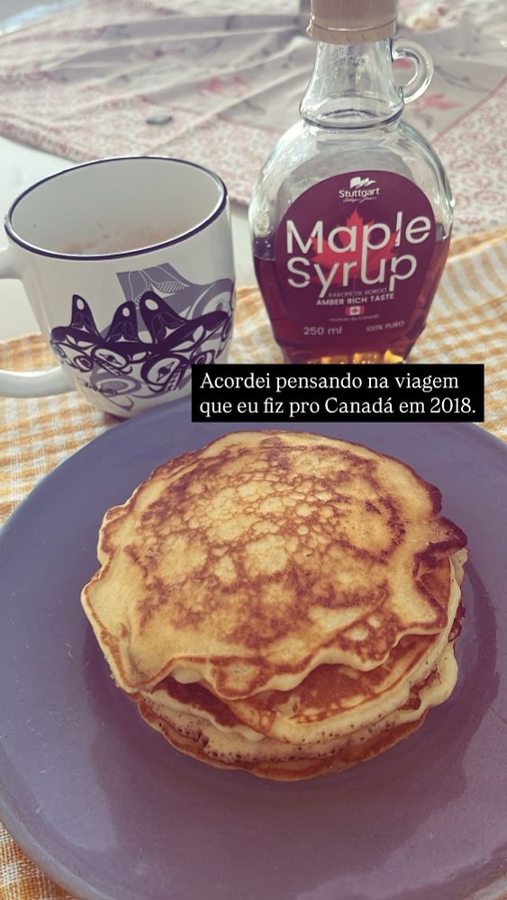 Foto de uma mesa de café da manhã coberta por uma toalha xadrez amarela, com um prato azul contendo uma pilha de panquecas americanas, uma caneca com desenhos de orcas no estilo indígena haida, e um vidro de xarope de maple. Tem um bloco de texto que diz “acordei pensando na viagem que eu fiz pro Canadá em 2018”.