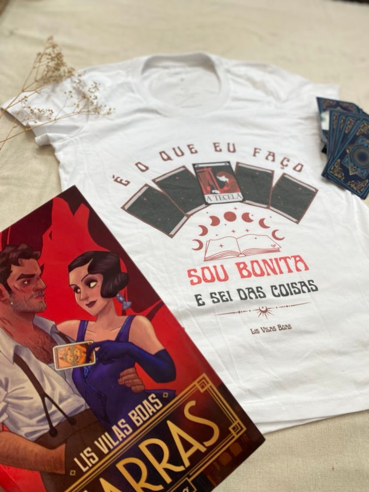 Fotografia do livro Garras ao lado de uma camiseta branca com a estampa em que está escrito “é o que eu faço, sou bonita e sei das coisas” que mostra cartas de baralho sobre um livro aberto com as fases da lua.