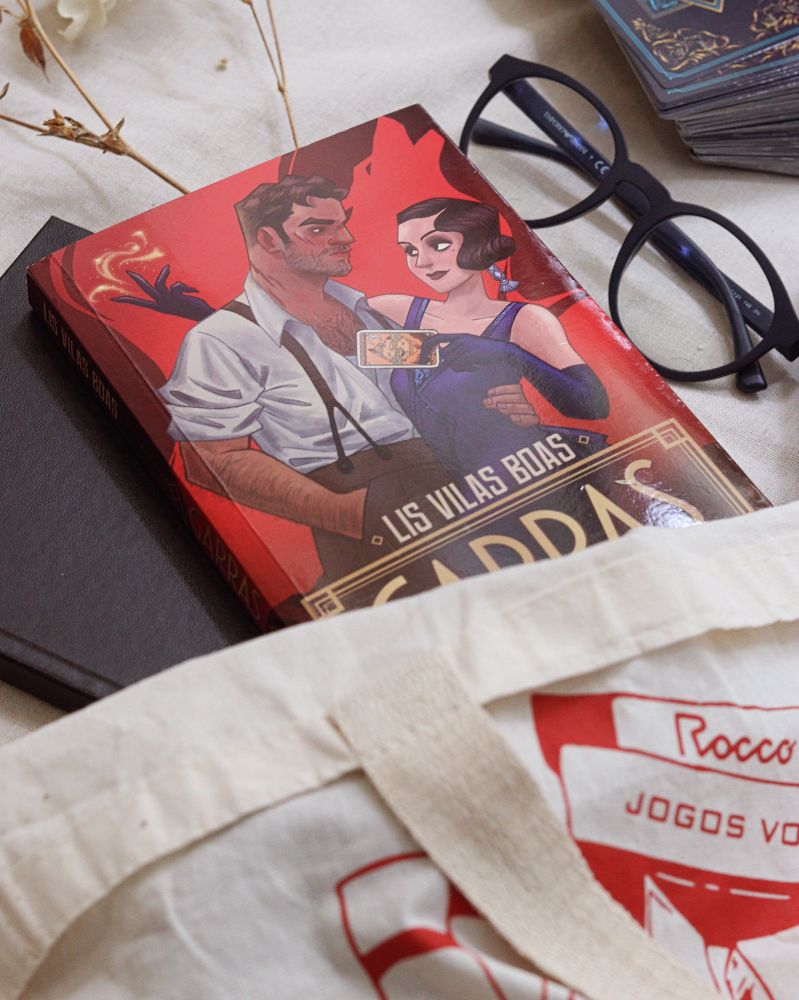 Fotografia de uma sacola de algodão com a logo da editora Rocco em vermelho meio aberta. Da sacola estão saindo um caderno de capa de couro preta, o livro Garras, óculos de armação azul, um galho de flores secas e um baralho de tarô empilhado.