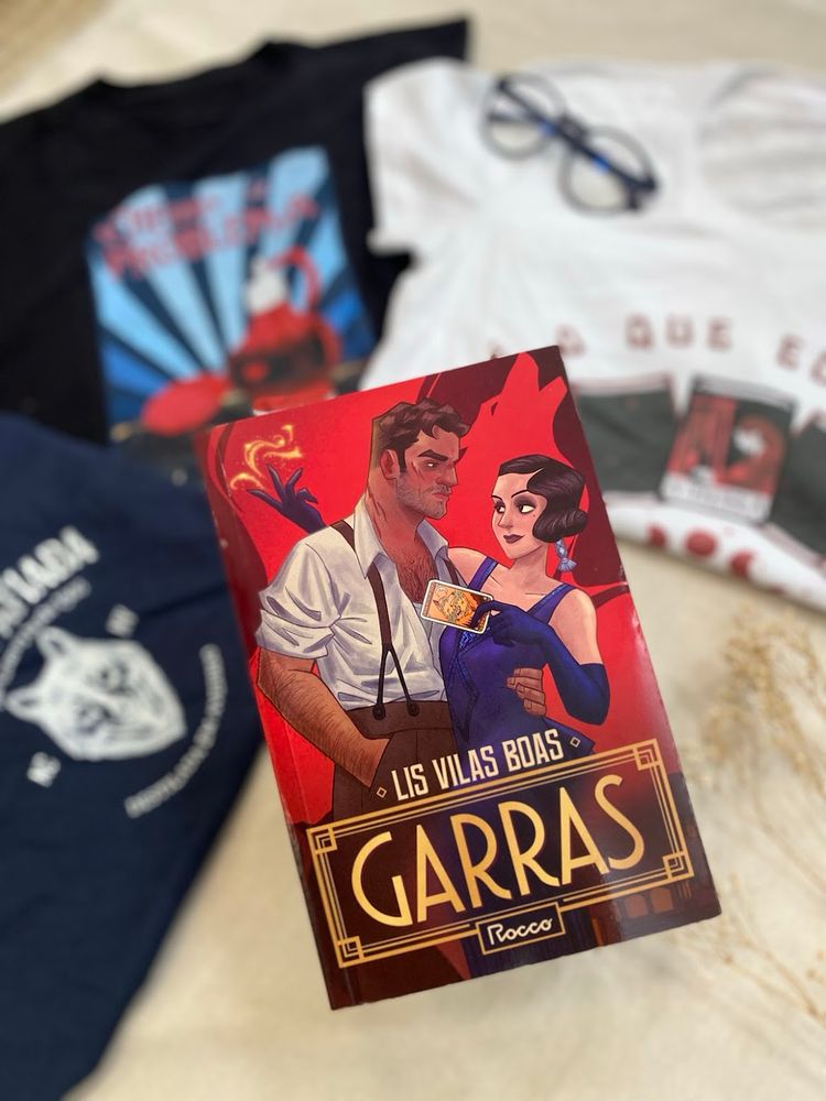 Fotografia do livro Garras em primeiro plano com três camisetas estampas das ao fundo, desfocadas.