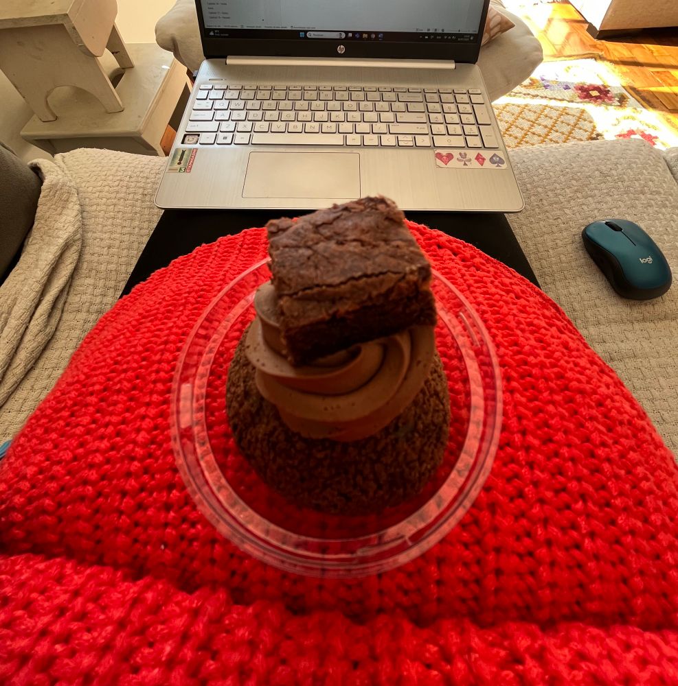 Fotografia quadrada de uma barriga de gestante com um choux de brownie apoiado em cima, por trás se vê um notebook aberto apoiado em pernas.