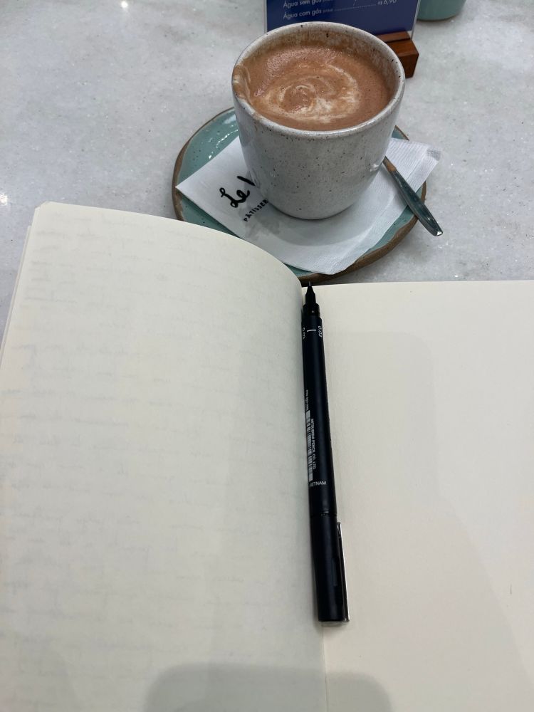 Fotografia de um caderno aberto com páginas em branco, com uma caneta por cima, ao lado de uma xícara de cerâmica com chocolate quente.