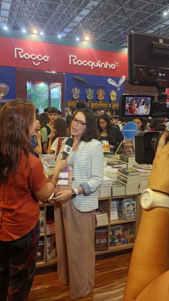 Fotografia da autora Lis Vilas Boas sendo entrevistada pela repórter Marina Araújo na frente de uma câmera no estande da editora Rocco na bienal