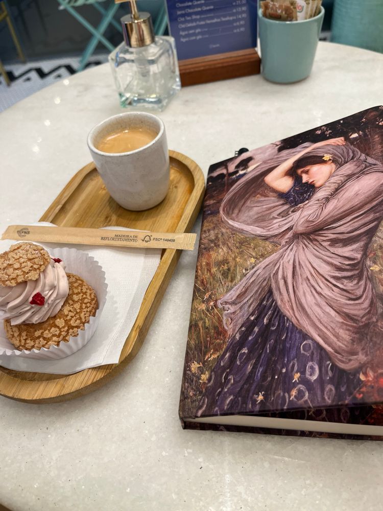 Fotografia de um caderno fechado com capa ilustrada de uma mulher de vestido azul e véu cinza, ao lado de uma bandeja oval de madeira contendo uma xícara pequena de café espresso descafeinado e um choux de framboesa.
