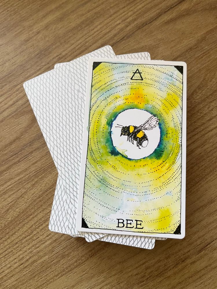 Fotografia de um baralho empilhado em cima da mesa com só uma carta virada para cima. A carta mostra um desenho que mistura aquarela e nanquim de uma abelha em voo, vista pela lateral, no centro de círculos pontilhados concêntricos e um fundo com tons de verde e amarelo.