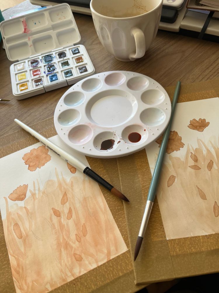 Fotografia de uma mesa com um godê branco com dois buraquinhos preenchidos por tinta de aquarela de tons marrons sobre dois cartões com artes florais em aquarela começados, e dois pincéis jogados sobre eles.