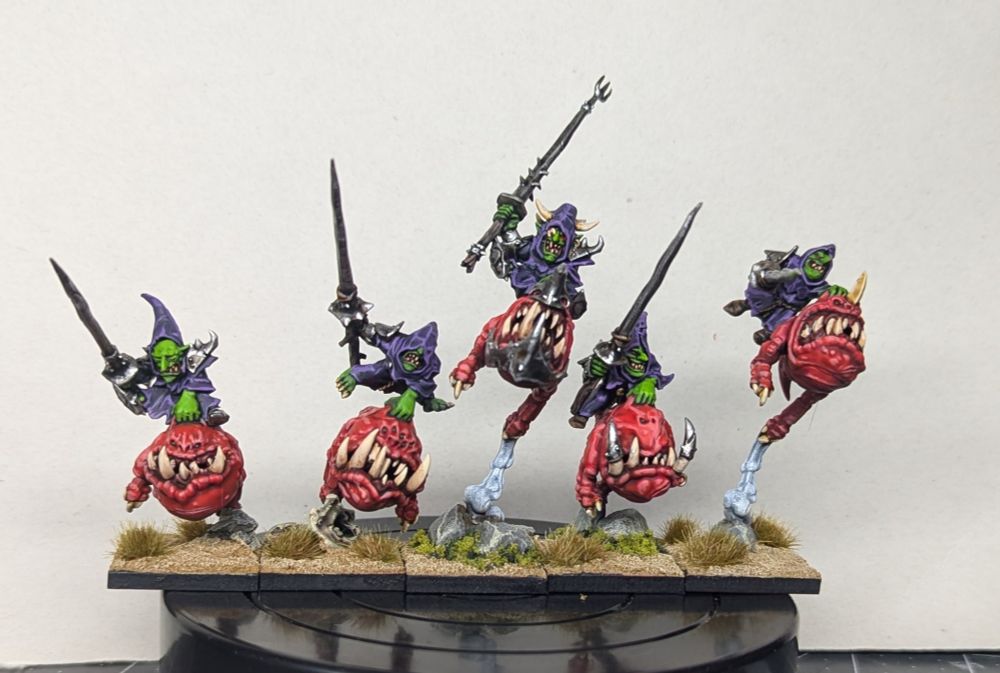 5 goblin squig riders with purple robes and bright green skin wielding lances. The squigs are bright red with stained ivory colored teeth and horns. 