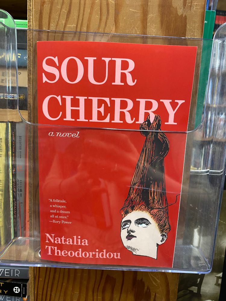 Red book cover with a woman’s head on it, title “Sour Cherry” by Natalia Theodoridou, seen in a bookstore